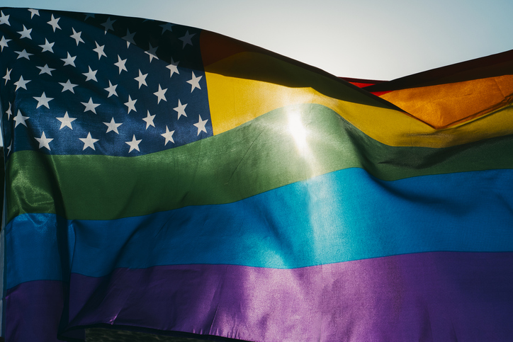 A rainbow US flag is waving on the sky, moved by the wind, with the sun in the background