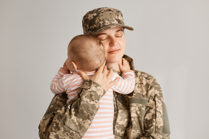 Happy young adult Caucasian military service woman soldier holding her infant baby with her eyes closed.